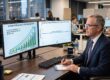An executive sitting at a desk with a large “AI” chart on one monitor and a flat productivity graph on another.