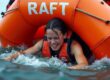 A life raft at sea labelled "RAFT" is helping a struggling swimmer.